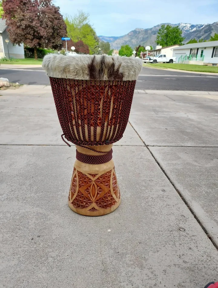Djembe with burgundy-and-black rope, a pale patterned hair-on rim, and deep geometric carving on a wooden base
